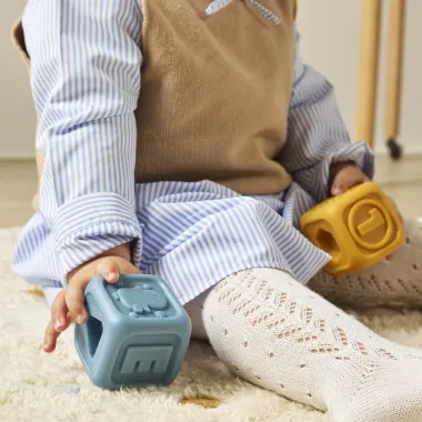 Kūdikis laiko Miniland minkštas kaladėles mėlynos ir geltonos spalvos.