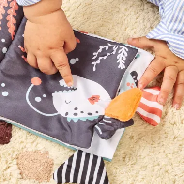 Mergaitė žaidžia su Miniland medžiagine knygele kūdikiams - su tekstūromis ir kontrastingais raštais.