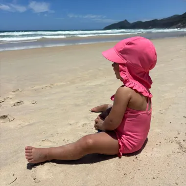 Mergaitė sėdi paplūdimyje su ružava Vanilla Copenhagen maudymosi kepuryte